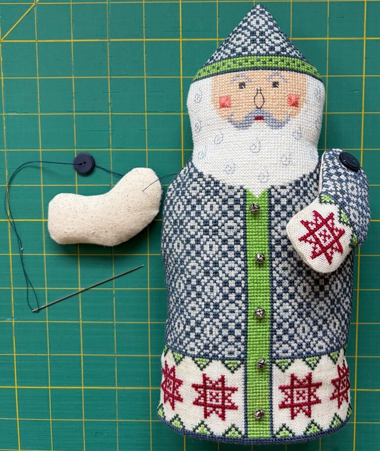 Santa with left arm attached, and right arm about to be pinned to body, with button strung on floss with needle.