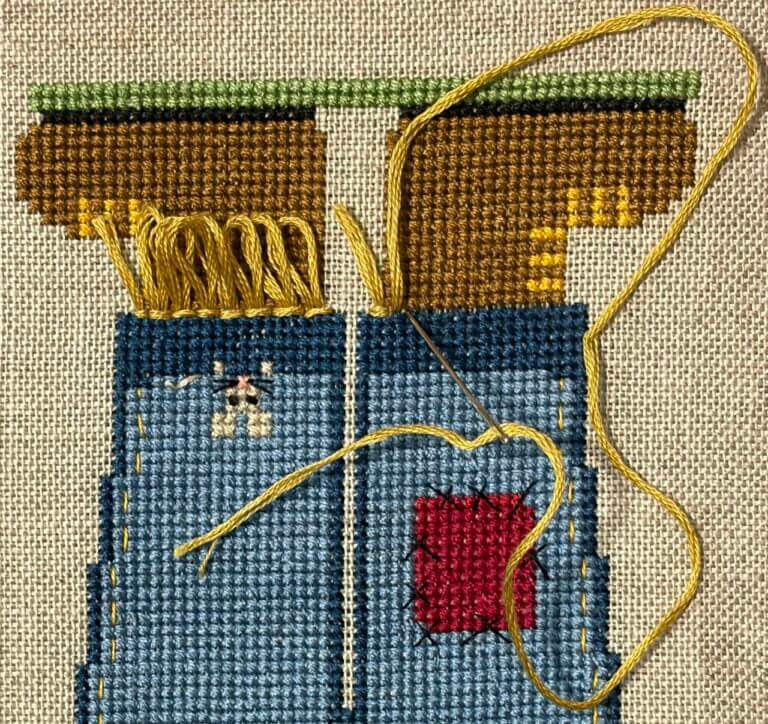 Close-up of blue cross-stitched pant legs with a needle forming golden-yellow loop stitches between the pants and boots