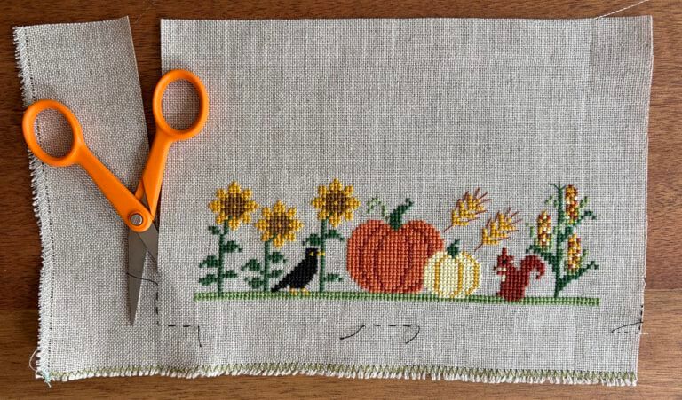 Orange-handled scissors trimming linen along a dashed running-stitch guideline beside a cross-stitched autumn band of sunflowers, pumpkins, corn, and a squirrel