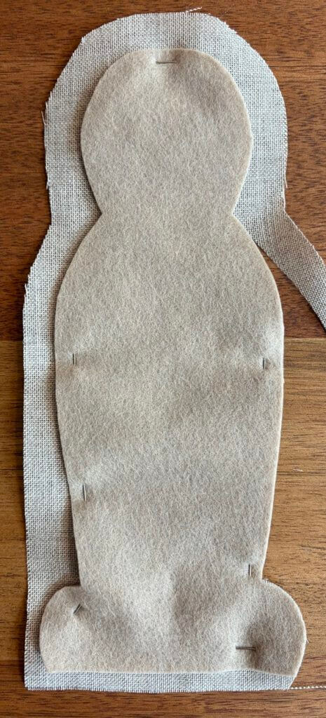 Tan felt body shape pinned onto a larger piece of cream linen, ready for the linen to be trimmed to the felt edge