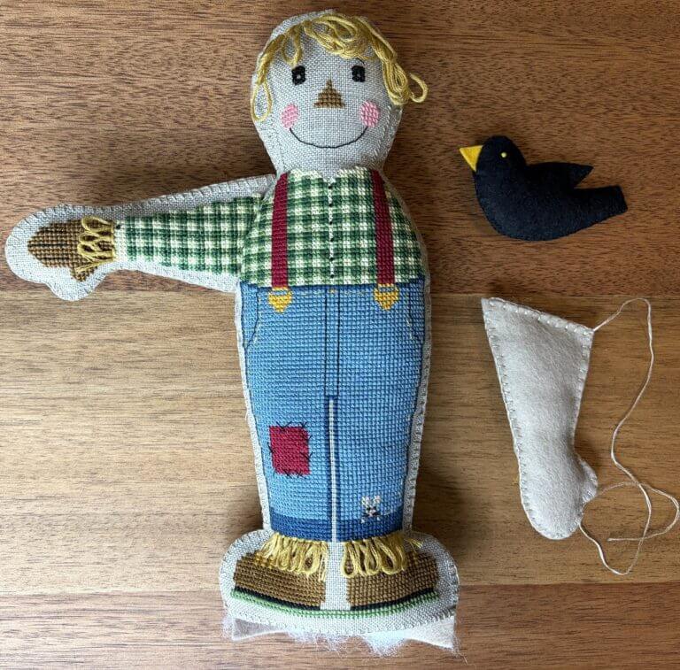 Partly assembled stuffed Harvest Henry with one plaid arm attached, the other arm and a small black crow piece laid beside him