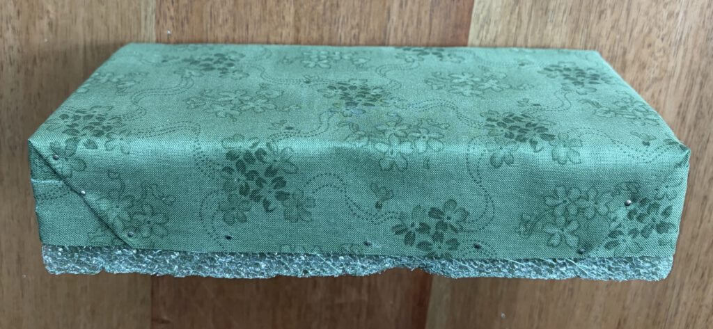 Rectangular foam block partly wrapped in green floral fabric with gift-folded corners, pinned along the lower edge where white foam still shows